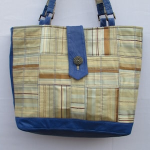 May include: A patchwork tote bag with a blue leather base, trim, and handles. The main body features a plaid pattern in shades of gold, tan, and light blue. A blue leather flap with a wooden button secures the bag's closure.