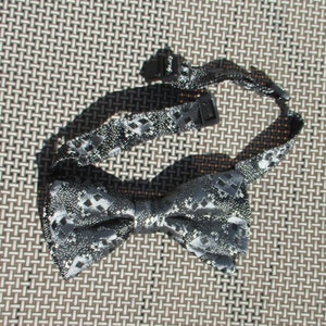 May include: A silver and gray bow tie with a geometric pattern. The bow tie is made of a textured fabric and has a shiny finish.