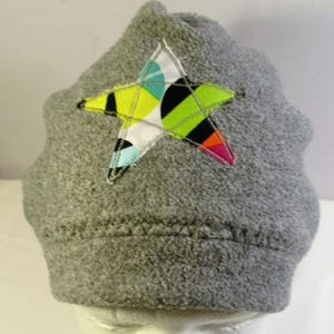 May include: A gray fleece beanie featuring a large, multi-colored star appliqué on the front. The star is patterned with geometric shapes in bright lime green, black, white, orange, pink, and light blue, secured with visible zigzag stitching.