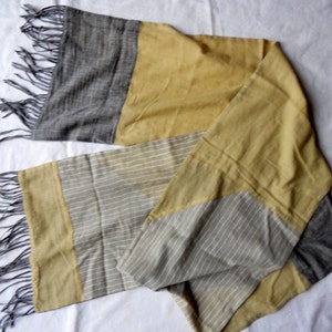 May include: A rectangular scarf with a color block design. The scarf features sections of gray, mustard yellow, and a light gray with white stripes. The ends have gray fringe. The scarf is made of a soft, lightweight fabric.