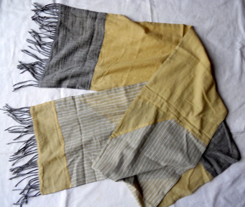 May include: A rectangular scarf with a color block design. The scarf features sections of gray, mustard yellow, and a light gray with white stripes. The ends have gray fringe. The scarf is made of a soft, lightweight fabric.