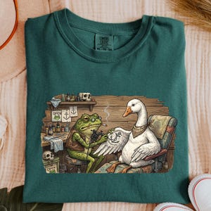 May include: Teal Comfort Colors t-shirt featuring a whimsical illustration of a frog tattooing a goose. The frog wears glasses and a vest, while the goose has a bandana. The tattoo reads "Adventure Co."