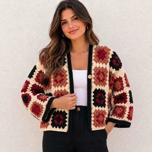 May include: A long-sleeve crochet cardigan featuring a granny square pattern in cream, black, dark red, and brown. The open-front garment has a single wooden button closure at the neckline and black trim along the edges, worn over a white top.
