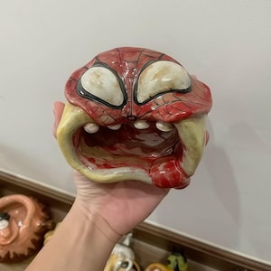 Könnte beinhalten: Eine Keramikschale in Form einer Spider-Man-Maske mit einem grotesken, geöffneten Mund. Die Schale hat ein rotes und schwarzes Netzmuster, große weiße Augen und gelbe und rote Akzente. Das Innere ist dunkelrot gefärbt.