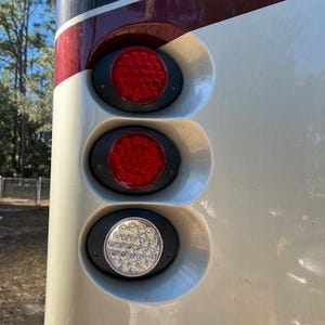 May include: Close-up of a vehicle's rear lights. Three oval-shaped lights are visible, with the top two emitting red light and the bottom one emitting white light. The lights are set against a cream-colored surface with a maroon stripe.