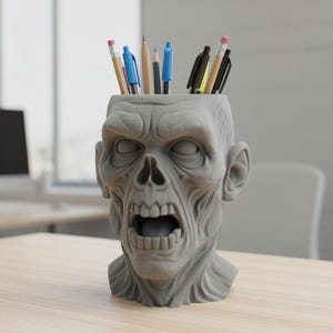 Peut inclure: Un organiseur de bureau en forme de tête de zombie grise rempli de crayons et de stylos. La tête de zombie à bouche ouverte est faite d'un matériau texturé et repose sur un bureau en bois. Les crayons et les stylos sont de couleurs variées.