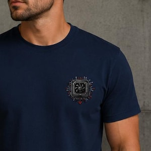 Könnte beinhalten: Ein dunkelblaues T-Shirt mit kurzen Ärmeln und einer kleinen, mittig platzierten Grafik auf der Brust. Die Grafik zeigt eine detaillierte, quadratische Computerprozessoreinheit mit einem zentralen Lüfter und roten sowie silbernen Details.