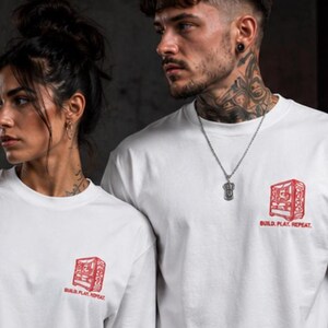 Könnte beinhalten: Ein weißes T-Shirt mit kurzen Ärmeln und einer kleinen roten Grafik auf der linken Brust, die einen Computerturm darstellt, mit dem Text "BUILD. PLAY. REPEAT.", der in Rot unter der Illustration gedruckt ist. Das Shirt hat einen Rundhalsausschnitt.