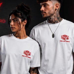 Könnte beinhalten: Ein weißes kurzärmeliges T-Shirt mit einer kleinen roten Grafik auf der linken Brust. Die Grafik zeigt einen Würfel mit dem darunter in Rot gedruckten Text "POWER. FOCUS. DOMINATE.". Das T-Shirt hat einen Rundhalsausschnitt.
