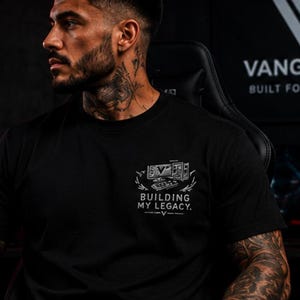 Könnte beinhalten: Ein schwarzes kurzärmeliges T-Shirt mit einer weißen Grafik auf der Brust. Die Grafik zeigt ein Computer-Setup mit Monitor und Tastatur, begleitet von dem Text "BUILDING MY LEGACY." und einem kleinen "V"-Logo, das unter dem Text zentriert ist.