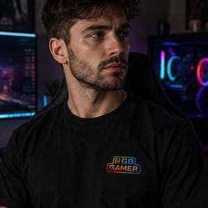 Peut inclure: Un t-shirt noir à manches courtes avec un petit motif rectangulaire multicolore sur la poitrine gauche. Le motif affiche le texte "RGB GAMER" dans une police stylisée avec un dégradé de couleurs arc-en-ciel sur un fond sombre.