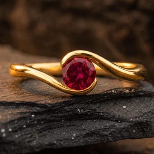 Pode incluir: Um anel de ouro com uma única pedra preciosa vermelha vibrante de corte redondo cravada no centro. A banda tem um acabamento liso e polido, curvando-se em torno da pedra num estilo cruzado, criando um design fluido e contínuo.