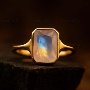 May include: A gold ring featuring a rectangular, faceted moonstone with a visible blue and yellow iridescent sheen. The gemstone is set in a smooth, polished gold bezel setting that transitions into a simple, slender gold band.