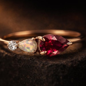 May include: A rose gold ring featuring three gemstones. A round, clear gemstone is on the left, followed by a pear-shaped opal, and a large, pear-shaped, deep red gemstone on the right. The ring is set against a dark background.