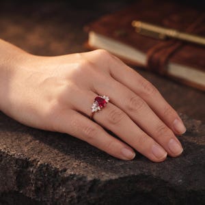 K&ouml;nnte beinhalten: Ein goldener Ring mit einem zentralen, oval geschliffenen, tiefroten Edelstein, der an beiden Seiten von kleinen, wei&szlig; geschliffenen Marquise-Steinen flankiert wird. Das Band ist d&uuml;nn und metallisch und hat eine polierte Oberfl&auml;che.