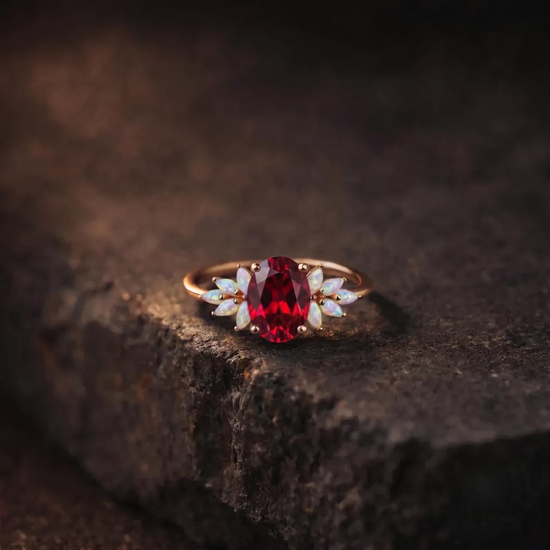 K&ouml;nnte beinhalten: Ein goldener Ring mit einem zentralen, oval geschliffenen, tiefroten Edelstein, der auf jeder Seite von drei kleinen, marquise-geschliffenen, irisierenden wei&szlig;en Opalen flankiert wird. Die Ringschiene ist schmal und poliert.