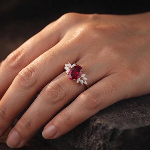 K&ouml;nnte beinhalten: Ein goldener Ring mit einem gro&szlig;en, oval geschliffenen, tiefroten Edelstein in der Mitte, flankiert von drei kleinen, marquise-geschliffenen, wei&szlig;en opalisierenden Steinen auf jeder Seite. Der Ring ist schmal und metallisch mit einer Krappenfassung.