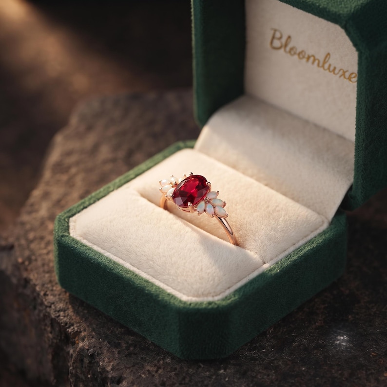 K&ouml;nnte beinhalten: Ein goldener Ring mit einem zentralen, oval geschliffenen, tiefroten Edelstein, der von kleinen wei&szlig;en Opalen in blattartiger Anordnung flankiert wird. Der Ring liegt in einer dunkelgr&uuml;nen Samtschatulle mit dem goldenen Schriftzug Bloomluxe.