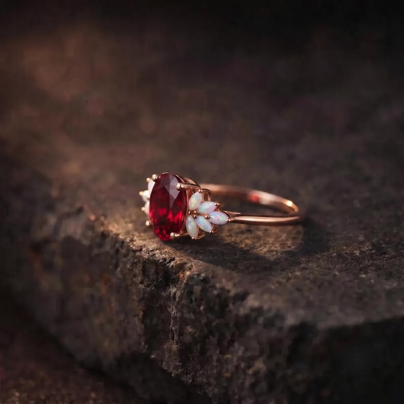 K&ouml;nnte beinhalten: Ein Ring aus Ros&eacute;gold mit einem zentralen, oval geschliffenen, tiefroten Edelstein. Die Fassung ist auf beiden Seiten mit jeweils drei kleinen, marquise-geschliffenen wei&szlig;en Opalen in blattartiger Anordnung verziert.