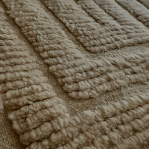 Peut inclure: Gros plan sur un tapis beige texturé avec un motif géométrique. Le tapis présente un motif à poils bouclés en relief, créant une surface douce et tactile. La couleur neutre et la texture épaisse suggèrent une ambiance chaleureuse.