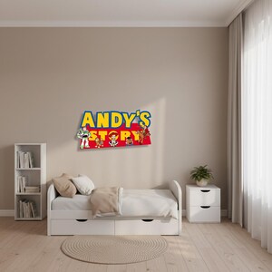 K&ouml;nnte beinhalten: Eine farbenfrohe Wanddekoration mit dem Text "Andy's Story" in Gelb und Blau, mit Zeichentrickfiguren. Das Schild befindet sich &uuml;ber einem wei&szlig;en Bett mit einer beigen Decke und Kissen, was auf ein Kinderzimmer-Thema hindeutet.