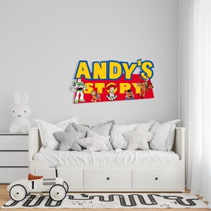 K&ouml;nnte beinhalten: Wanddekoration mit dem Text "ANDY'S STORY" in Gelb und Blau, mit Charakterillustrationen. &Uuml;ber einem wei&szlig;en Bett mit grauen sternf&ouml;rmigen Kissen. Ein wei&szlig;es Spielzeugauto und eine Hasenlampe befinden sich im Vordergrund.