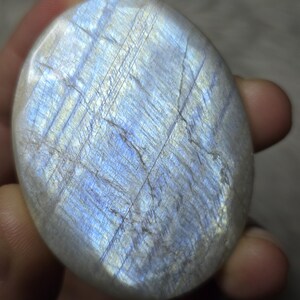 May include: A polished, oval-shaped moonstone with a pearlescent sheen. The stone displays a play of color, with flashes of blue and white across its surface. The gemstone is held in a hand, showcasing its smooth texture and natural beauty.
