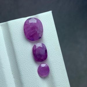 May include: Three faceted, oval and round, deep pink gemstones. The largest is an oval shape, the middle one is also oval, and the smallest is round. They are displayed on a white surface with a dark gray background.