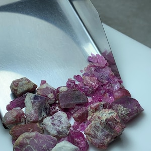 Könnte beinhalten: Eine Sammlung roher, ungeschliffener Rubin-Edelsteine in verschiedenen Rosa- und Rottönen. Die Edelsteine werden auf einer weißen Oberfläche präsentiert, wobei einige aus einer silbernen Metallschale herausfallen. Die rohen Steine haben ein natürliches, unpoliertes Aussehen.