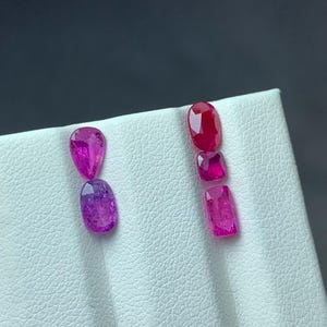 Pode incluir: Uma variedade de pedras preciosas polidas em tons de rosa e vermelho. As gemas são exibidas em uma superfície branca, com um fundo escuro. As formas incluem cortes ovais, em forma de pêra e retangulares. As gemas são provavelmente rubis.