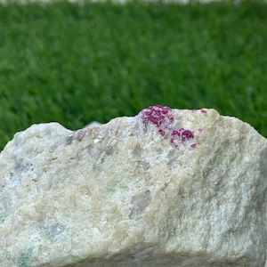 May include: A raw, rough mineral specimen featuring a light beige, textured stone matrix with small, embedded clusters of vibrant, deep magenta-colored crystals concentrated along the top edge of the irregular, jagged rock formation.