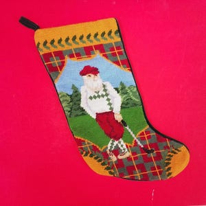 May include: Needlepoint Christmas stocking featuring Santa Claus playing golf. The stocking has a yellow top with green trim, a plaid pattern, and a scene of Santa on a golf course. The background is red.