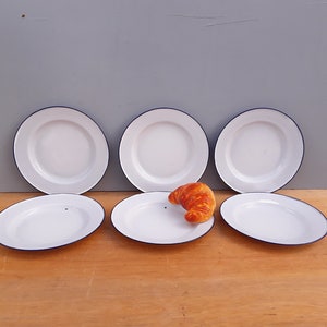 May include: Six white enamel plates with a blue rim. The plates are arranged in a semi-circle on a wooden surface. A croissant is sitting on one of the plates.