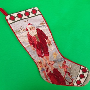May include: A red and white Christmas stocking with an embroidered image of Santa Claus on a beach with other figures. The stocking has a green and white diamond pattern at the top and bottom.