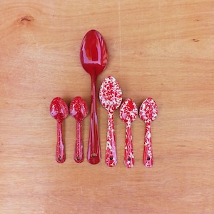 May include: A set of six spoons arranged in a row. One large solid red spoon is positioned next to five smaller spoons featuring a red and white speckled enamel pattern. Each spoon has a rounded bowl and a handle with a small hole at the end.