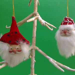 May include: Two Christmas ornaments shaped like Santa Claus faces. One Santa has a red hat with black polka dots, the other has a red and white hat. Both have white beards and rosy cheeks, hanging from gold strings.