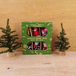 May include: A green box labeled "A Shiny Brite Product" contains six miniature musical dwarf band figures. Two small artificial Christmas trees flank the box on a wooden surface. The box has a decorative border and text.