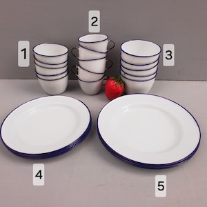May include: A collection of white enamelware dishes with a blue rim. The set includes several cups, plates, and a strawberry. The cups have black handles. The plates are oval-shaped.