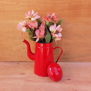May include: A red enamel teapot with a lid, filled with pink flowers. The teapot has a spout and a handle.