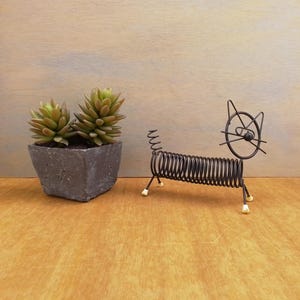 May include: A dark brown wire sculpture shaped like a cat with a coiled spring body, a circular head, and small white-tipped feet, positioned next to a square gray planter containing two green succulent plants with pointed leaves.