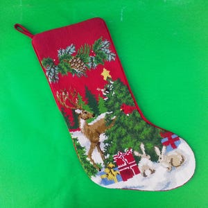 May include: A red needlepoint Christmas stocking featuring a detailed scene of a deer, a decorated pine tree, two rabbits, and wrapped gifts on a snowy ground. The top section displays green holly leaves and a pinecone, with a red hanging loop.
