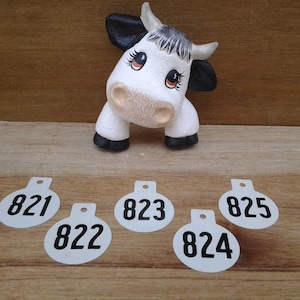 May include: Five white round plastic tags with black numbers on them. The numbers are 821, 822, 823, 824, and 825. A white ceramic cow figurine is in the background.