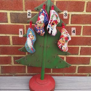 May include: A green metal Christmas tree with a red base holds four small Christmas stockings. The stockings are decorated with images of Santa Claus and other holiday designs. The tree is set against a red brick wall.
