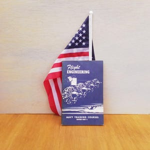 May include: A vintage navy blue book titled "Flight Engineering" with illustrations and the text "Navy Training Courses." An American flag is positioned behind the book. The book and flag are on a wooden surface.