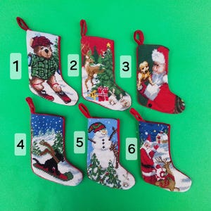 May include: Six Christmas stockings with red hanging loops. Each stocking features a different embroidered holiday scene, including a bear on skis, a reindeer, Santa Claus, and a snowman. The stockings are set against a bright green background.