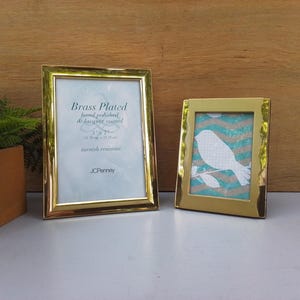 May include: Two brass-plated picture frames with a polished finish. The larger frame contains a paper insert with the text Brass Plated, hand polished & lacquer coated, 5 x 7, tarnish resistant, JCPenney. The smaller frame holds a bird graphic.