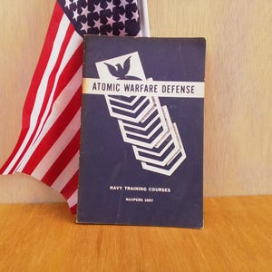 May include: A blue booklet with the title "Atomic Warfare Defense" and the text "Navy Training Courses NAVPERS 10097". A white dove with outstretched wings is above the title. The booklet is in front of a folded American flag.