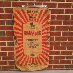 May include: Tan burlap feed sack with red lettering and graphics. The bag reads "WAYNE" in large letters, with "POULTRY AND LIVESTOCK FEEDS" below. The bag is marked "100 LBS. NET" and is hanging on a clear plastic hanger.