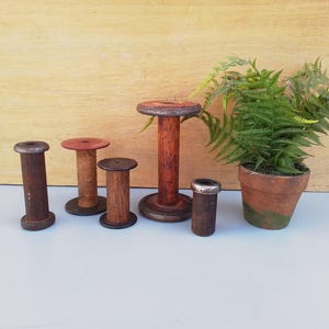 May include: A collection of vintage wooden spools in varying sizes and shades of brown, with a small potted fern. The spools have a rustic, aged appearance, suggesting they are antique or reclaimed items. The fern is in a terracotta pot.