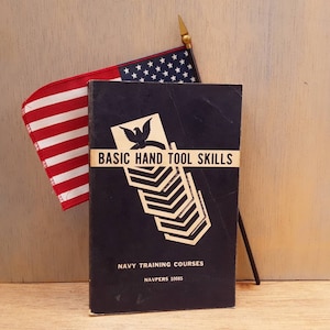 May include: A navy blue book titled "Basic Hand Tool Skills" with a white and black illustration of a bird and a series of chevron shapes. The book is part of the Navy Training Courses series and has the number NAVPERS 10085 printed on the cover. The book is sitting on a wooden surface with an American flag draped over it.